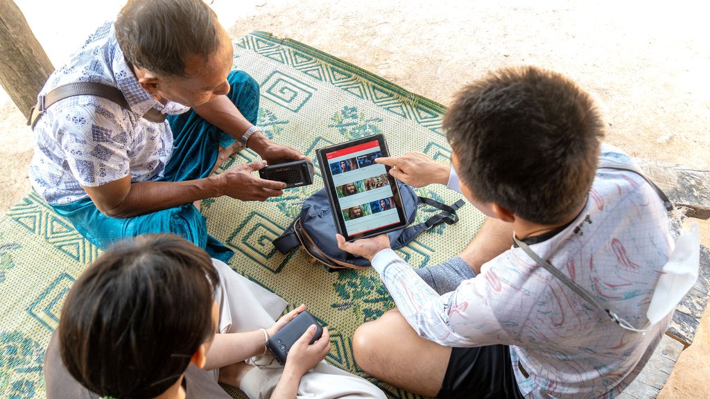 Young man showing tablet with Jesus Film app to two other people while sitting down on a mat